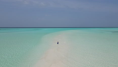 Young-girl-wearing-a-long-blue-dress-running-barefoot-on-a-surreal-white-sandbank-with-turquoise-waters-of-Indian-ocean,-Maldives-drone-shot-on-a-clear-sunny-day