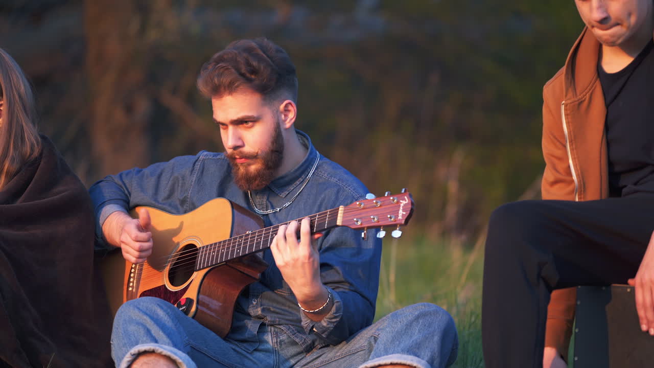 Group of friends spending time together in the nature. Guys playing guitar and singing songs sitting on the ground at sunset.