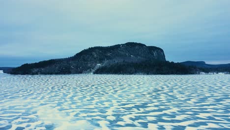 Tobogán-Aéreo-Sobre-Un-Lago-Congelado-Con-Patrones-Nevados-Más-Allá-De-Una-Montaña-Aislada-Con-Un-Acantilado-Empinado