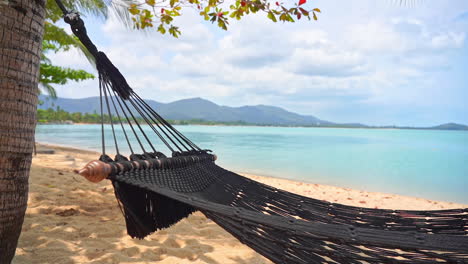 Close-up-A-robe-hammock-suspended-over-a-golden-sandy-beach-and-shoreline-sways-in-the-tropical-breeze