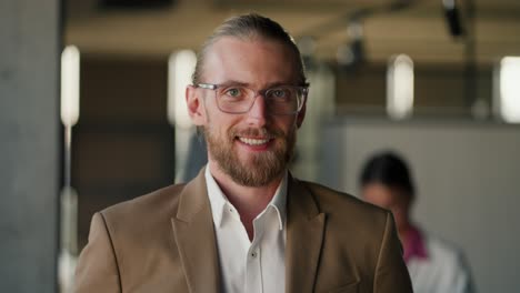 Portrait-of-a-blond-man-in-glasses-in-a-brown-jacket-and-white-shirt-who-looks-at-the-camera-and-smiles-against-the-of-a-modern-office.-Business-man-in-modern-office