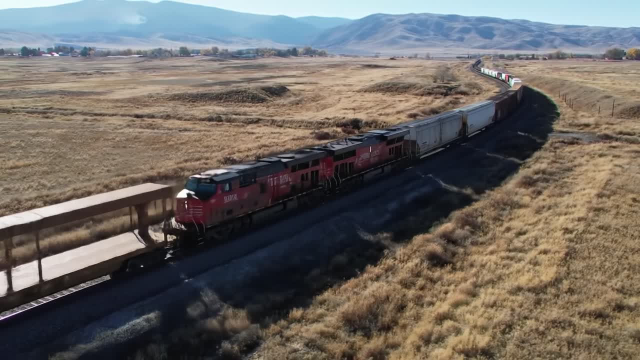 A long freight train moves through vast grasslands, surrounded by mountains and under a clear blue sky. The scenery captures the essence of autumn with changing foliage.