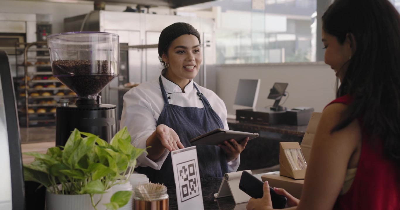 Chef in divisa che tocca il POS, cliente afroamericano che mostra il telefono per pagare, consegna di un pacchetto al bancone