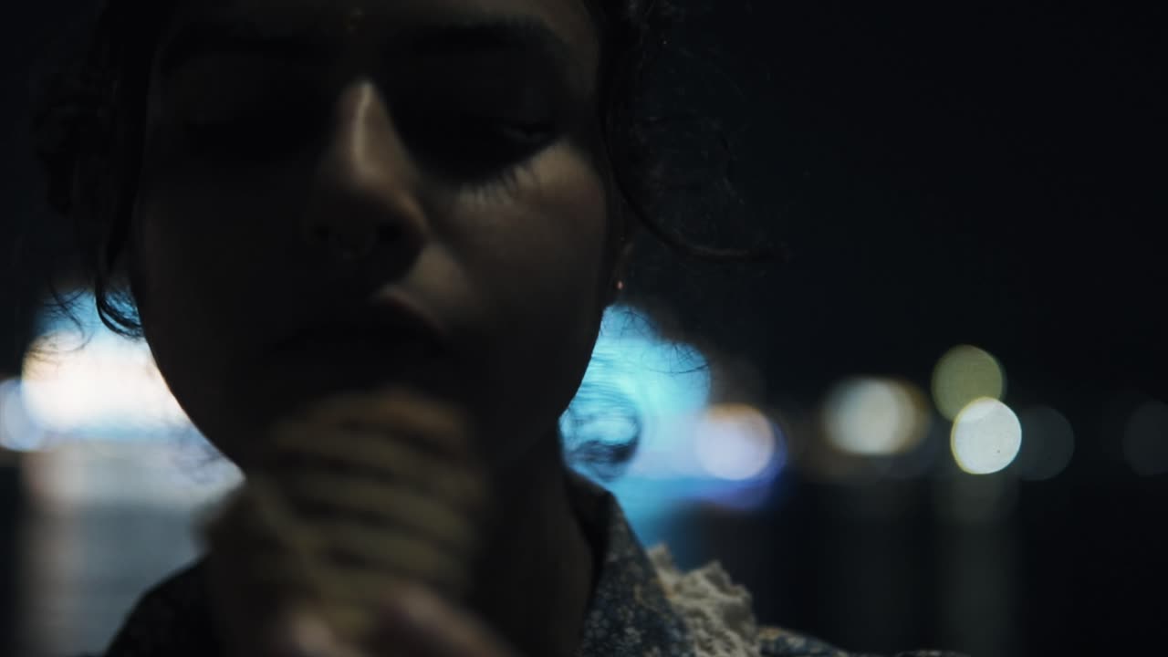 젊은 아름다운 인도 여성이 아이스크림을 먹고 이마에서 머리카락을 고, 배경에는 도시 불빛이 있습니다.