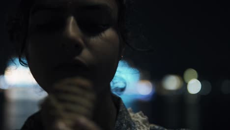 young beautiful indian woman eats ice cream and brushes her hair from her forehead, city lights in the background