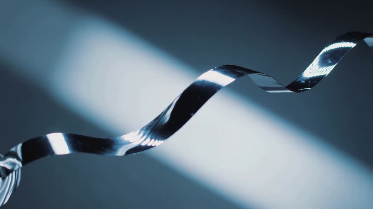 Rotating thin metal ribbon waving more in studio, motor turning diagonal beam revealing highlights