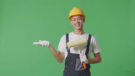 asian man painter wearing safety helmet smiling and pointing to side while standing in the green screen background studio
