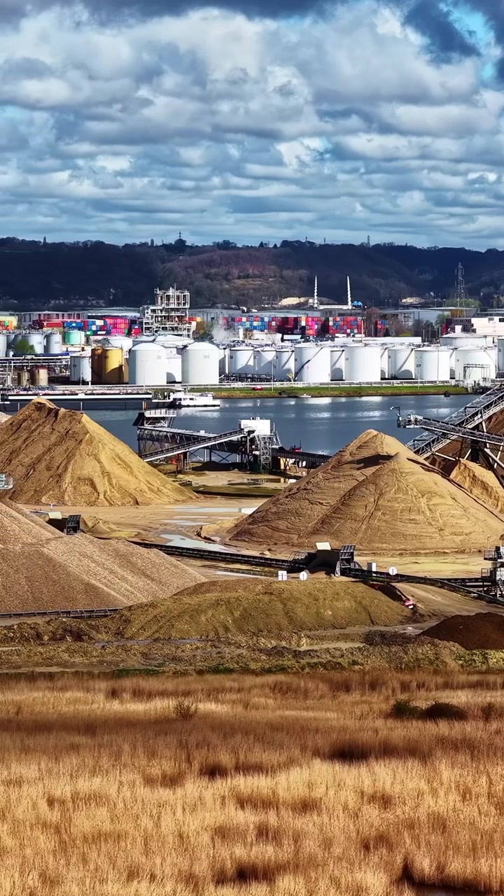 Sandhaufen und Lagertanks säumen eine Industrieanlage am Wasser unter einem dynamischen Himmel. Ideal für Umweltdokumentationen oder Projekte mit industriellem Thema.