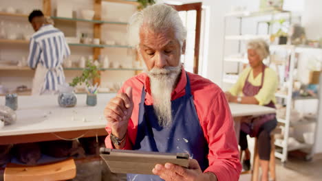 Alfarero-Birracial-Enfocado-Con-Barba-Larga-Usando-Tableta-En-Estudio-De-Cerámica,-Cámara-Lenta