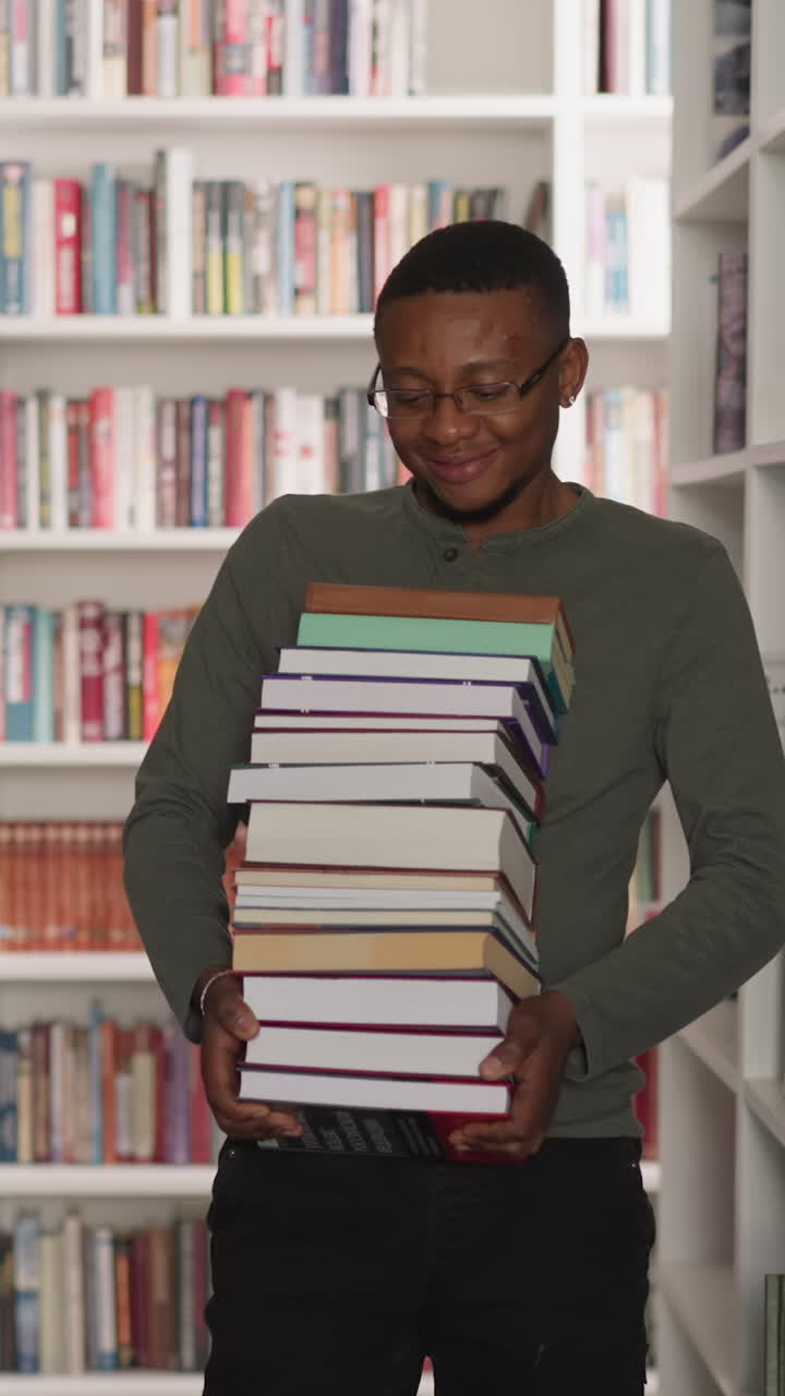 Emotional man carries book stack in library. African American librarian holds large literature pile walking between bookcases. Student in archive