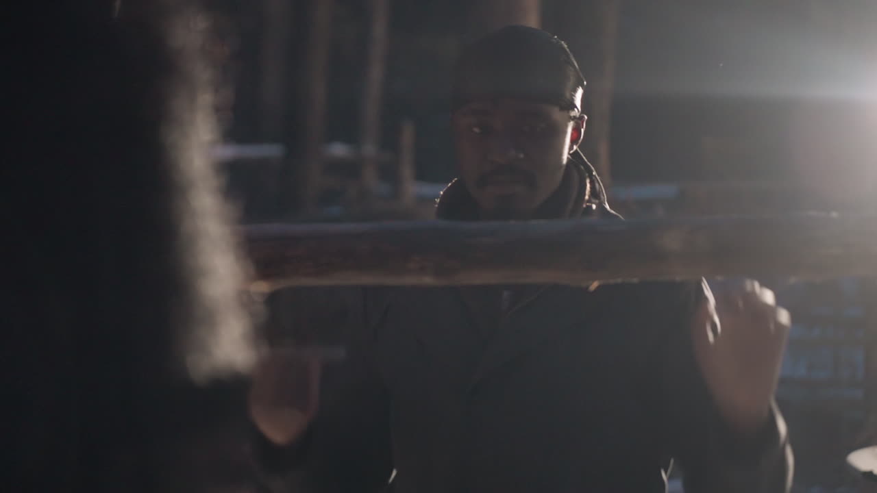 hombre negro levantando un tronco en un bosque al amanecer, aserradero accidentado de fondo, silueta fuerte y destello de contraluz, aire frío visible, expresión concentrada y trabajo manual que transmite determinación