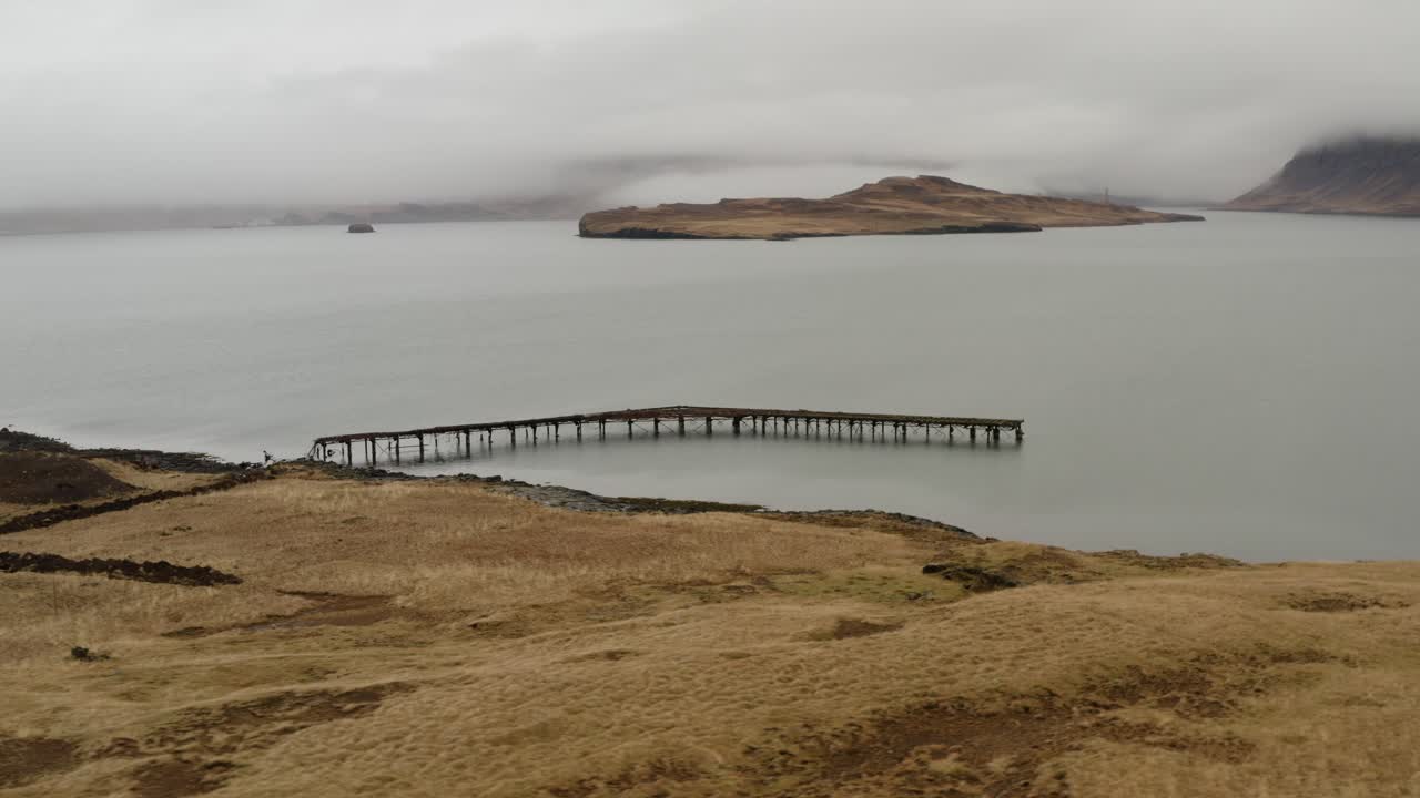 Vista rialzata di un molo arrugginito che si estende nella baia nebbiosa a Hvalfjörður, in Islanda, con erba secca in primo piano e un'isola lontana coperta di nebbia sull'acqua calma