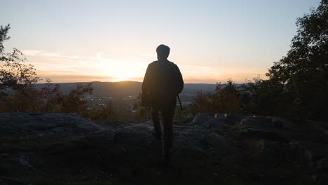 Young-man-finishes-a-hike-at-the-top-of-a-rocky-cliff-mountain-at-sunset-and-looks-out-at-a-beautiful-viewpoint