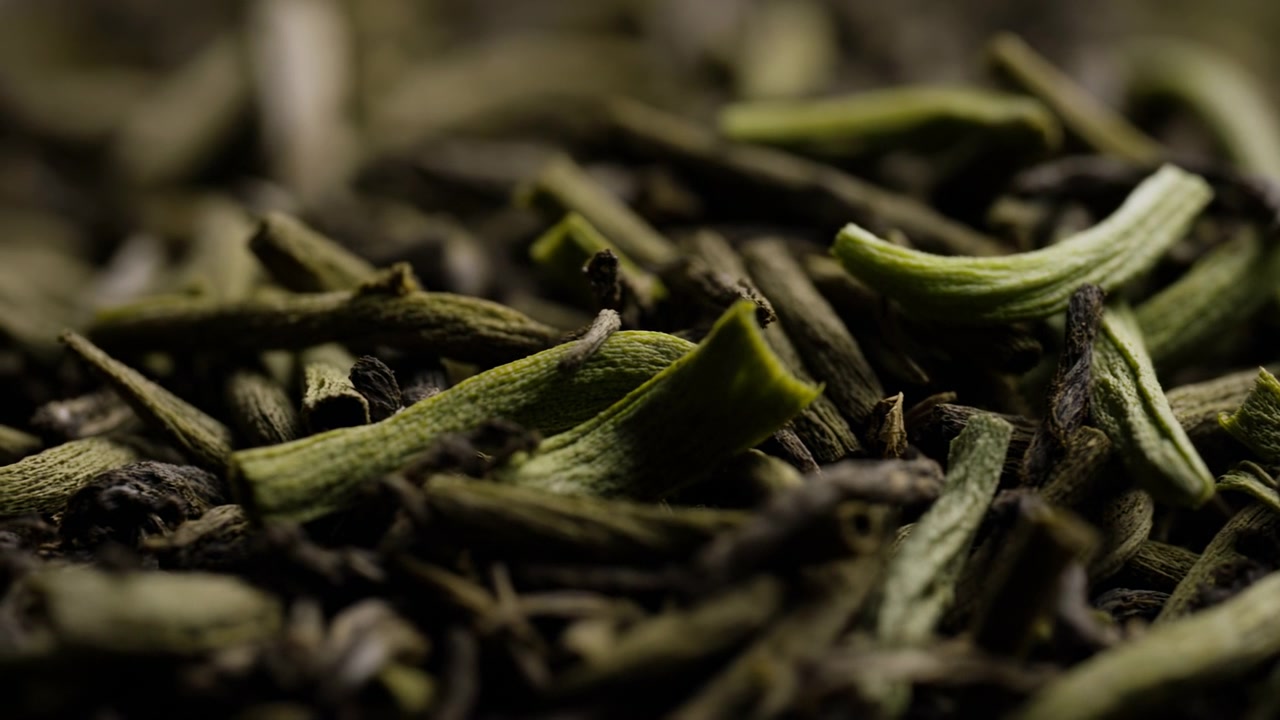 Primo piano dettagliato di foglie di tè verde essiccate disposte in una pila sovrapposta, mostrando segmenti arricciati e nervati, superfici opache fibrose, toni terrosi e occasionali punte scure e fragili