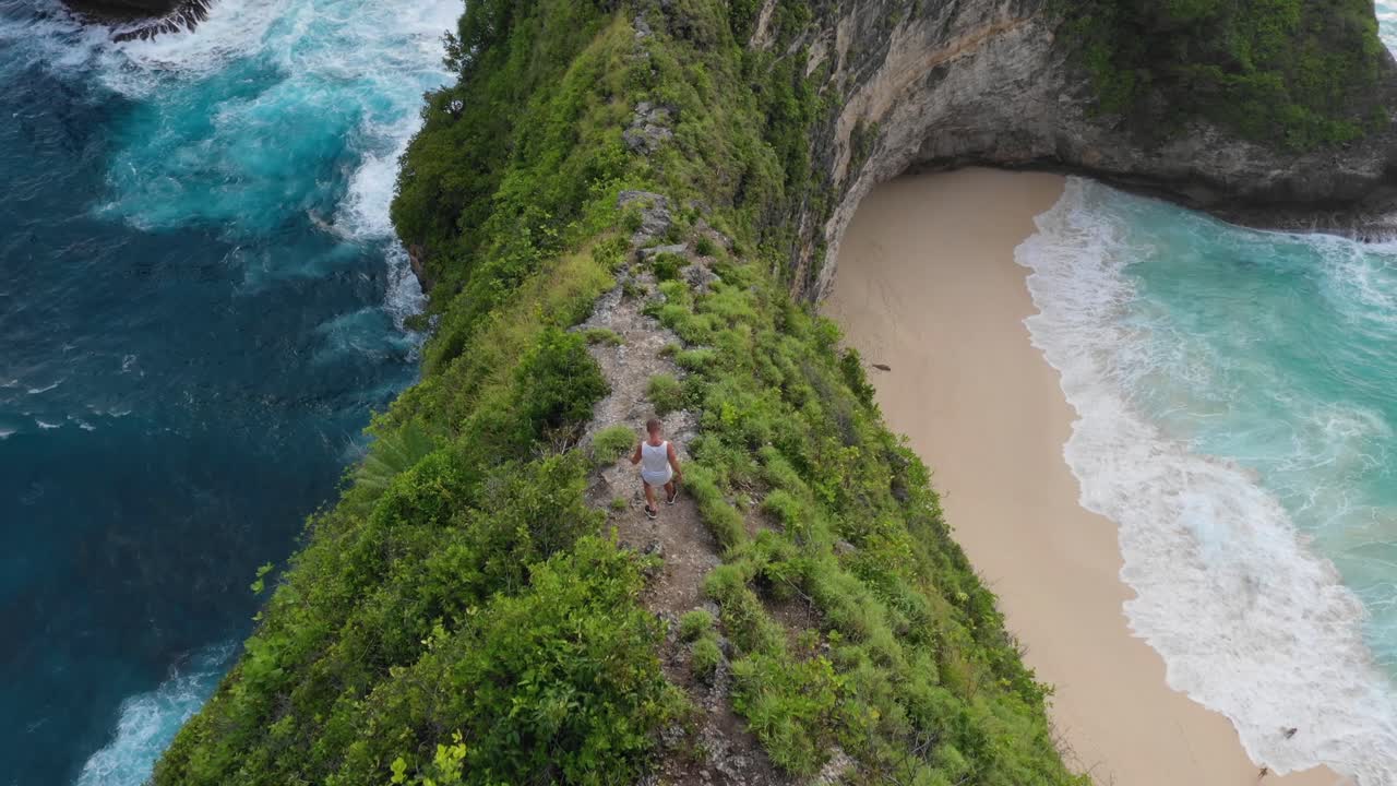 hombre caminando por un acantilado afilado con una playa idílica en el fondo, aérea, bali