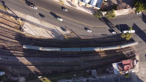 Aerial-overhead-shot-of-slow-train-arriving-Chacarita-Station-in-Buenos-Aires---Cars-driving-on-road-beside-during-sunset
