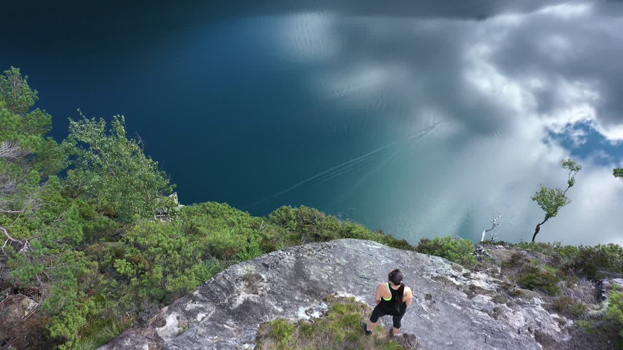 volando por encima de la cabeza de una mujer de pie en el borde de los acantilados - espectacular y peligroso cliff slottet en modalen noruega - mujer disfrutando del mirador y el fiordo con reflejos del cielo