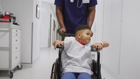african american male doctor walking with child patient sitting in wheelchair at hospital