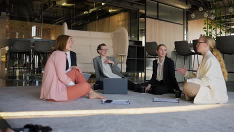 Zoom-in-approaching-a-group-of-confident-business-girls-in-business-uniforms-communicating-while-sitting-on-a-soft-carpet-in-a-modern-office