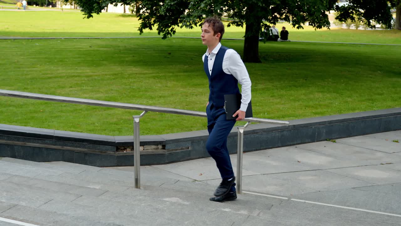 Young businessman in formal wear walking up outdoor stairs with a document case, symbolizing ambition, success, and career progression in a corporate urban environment, slow motion follow shot
