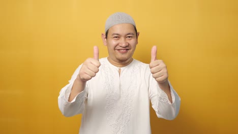 젊은 행복한 아시아 무슬림 남자가 엄지손가락을 올려 놓고 미소 짓고 행복한 얼굴을 보여 주고, 노란색 배경에 ok 표지판을 승인합니다.
