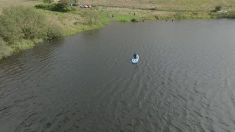 Tracking-Aerial-Shot-Of-A-Young-Woman-In-A-Wetsuit-Paddling-On-A-Stand-Up-Paddleboard-On-Loch-White