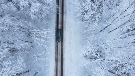 top-down view of a car driving through a snow-covered forest road, aerial drone hovering over village road in winter when a car passes by