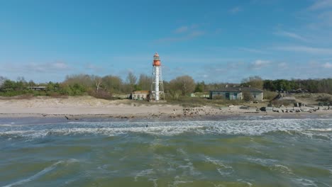 Aerial-establishing-view-of-white-colored-Pape-lighthouse,-Baltic-sea-coastline,-Latvia,-white-sand-beach,-large-waves-crashing,-sunny-day-with-clouds,-wide-ascending-drone-orbit-shot