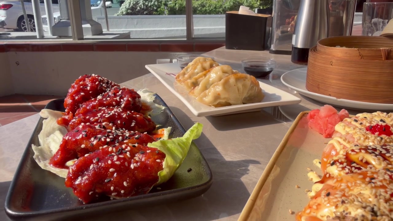 Close-up of glazed Korean fried chicken wings coated in red gochujang sauce and sesame seeds, plated with lettuce, alongside a plate of golden pan-fried dumplings, shot at a restaurant table.