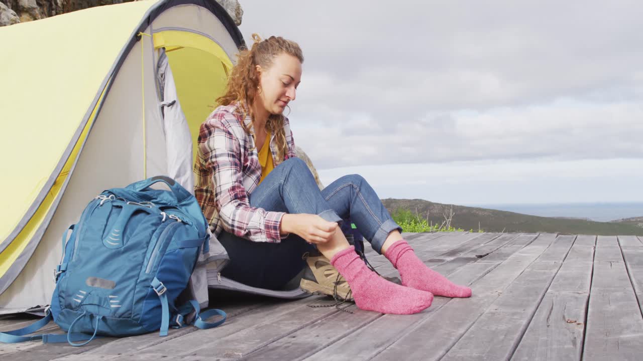 feliz mujer caucásica acampando, sentada fuera de la tienda poniéndose botas en el entorno rural de la montaña