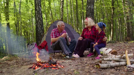 Senior-grandmother-grandfather-granddaughter-telling-funny-stories-fairy-tales-over-campfire-in-wood