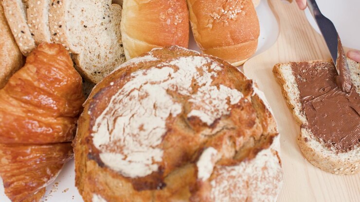 Close-up overhead shot of spreading chocolate cream on bread amidst various fresh bakery items.