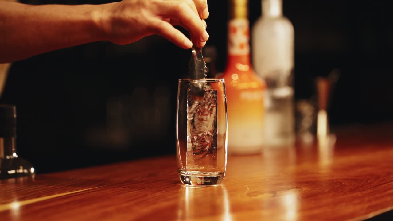 A close-up of a professional bartender's hand using tongs to carefully place a long, crystal-clear artisanal ice spear into a glass while preparing a premium cocktail