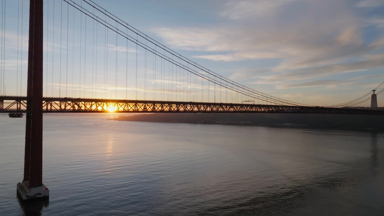 Video drone che riprende l'alba dietro l'iconico Ponte 25 de Abril di Lisbona mentre le auto lo attraversano sopra un fiume tranquillo, immerso in una splendida luce dorata e cieli drammatici in un'ampia ripresa panoramica