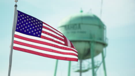 Bandera-Americana-En-Cámara-Lenta-Con-Menagha,-Torre-De-Agua-De-Minnesota-En-El-Fondo