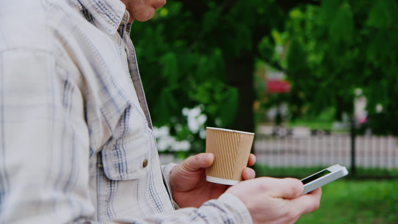 한 남자가 전화기와 커피 컵을 프레임에 들고 걷는다. 손만 보인다.