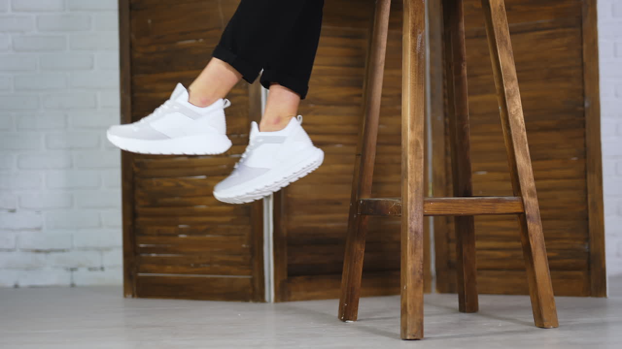 Unrecognizable woman sitting on a high wooden stool and waving feet. Model demonstrates white comfortable footwear.