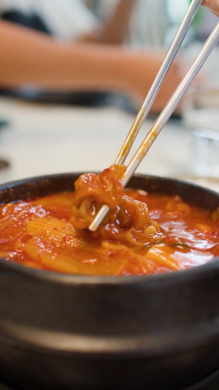 Bidikan close-up seseorang sedang makan sup kimchi pedas dengan sendok dan sumpit di restoran
