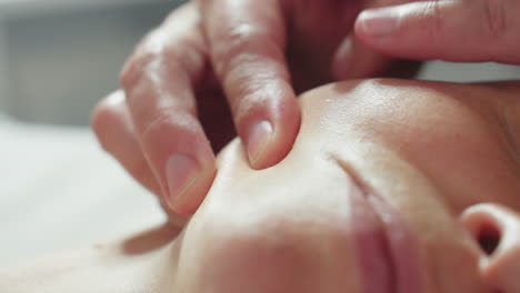 bodywork therapist cradles client side jawline with gentle fingertips applying circular massage strokes during serene facial under soft clinical lighting next to product trolley in treatment room