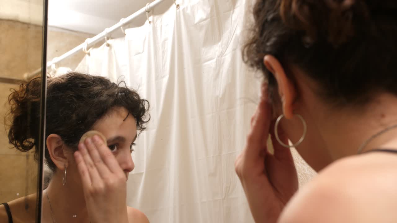 Giovane donna che si trucca con una spugnetta allo specchio del bagno