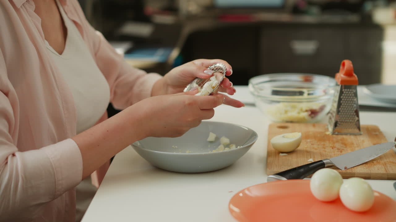 Culinary artist using small net egg smasher to crush boiled egg into bowl, working carefully with both hands in bright kitchen, surrounded by kitchen tools, knife, grater, and other ingredients