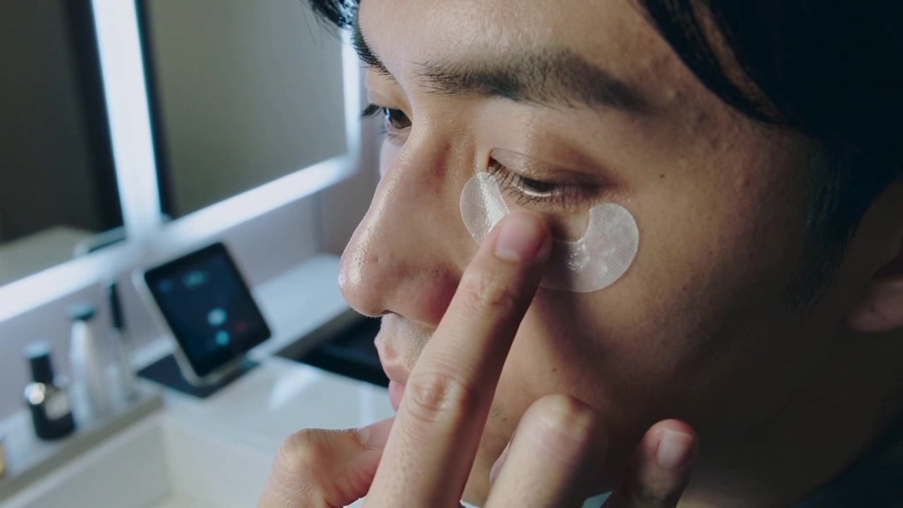 Applying Asian man wearing dark top smoothing clear gel patch under right eye at vanity, hydrating