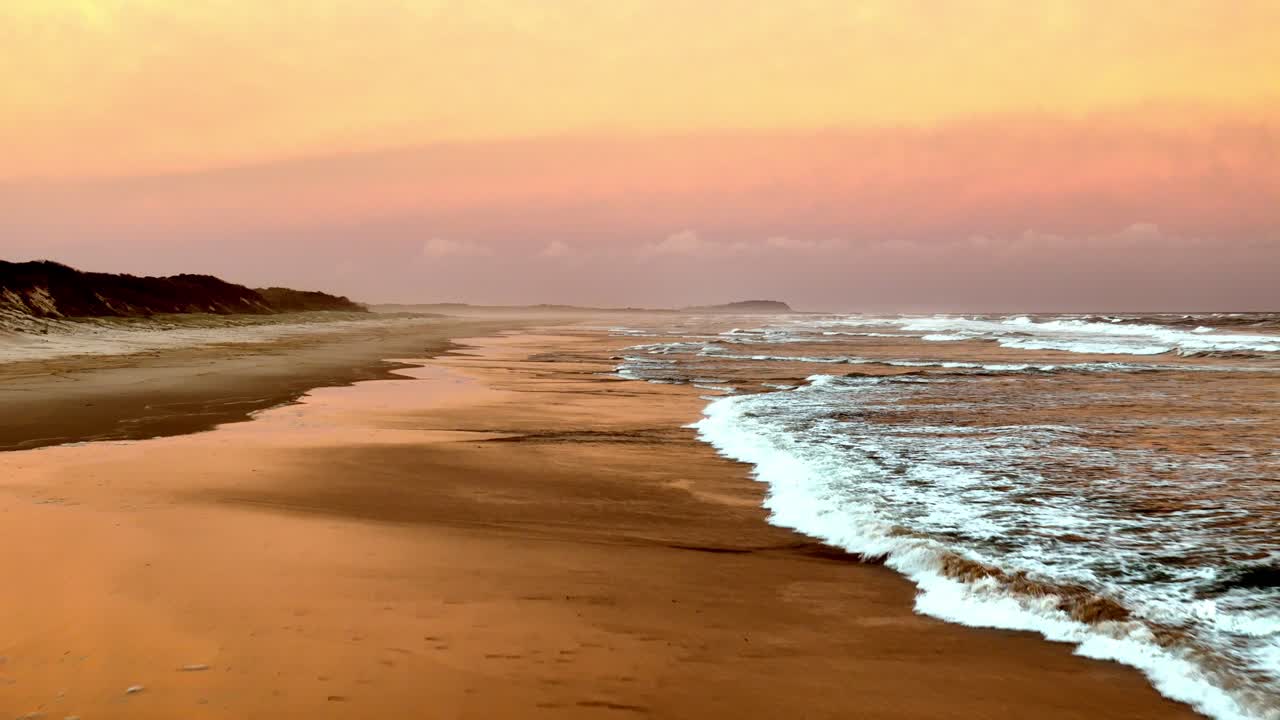 los suaves tonos pastel del amanecer reflejándose en la playa, las olas lavando suavemente sobre la arena bajo un cielo lleno de nubes