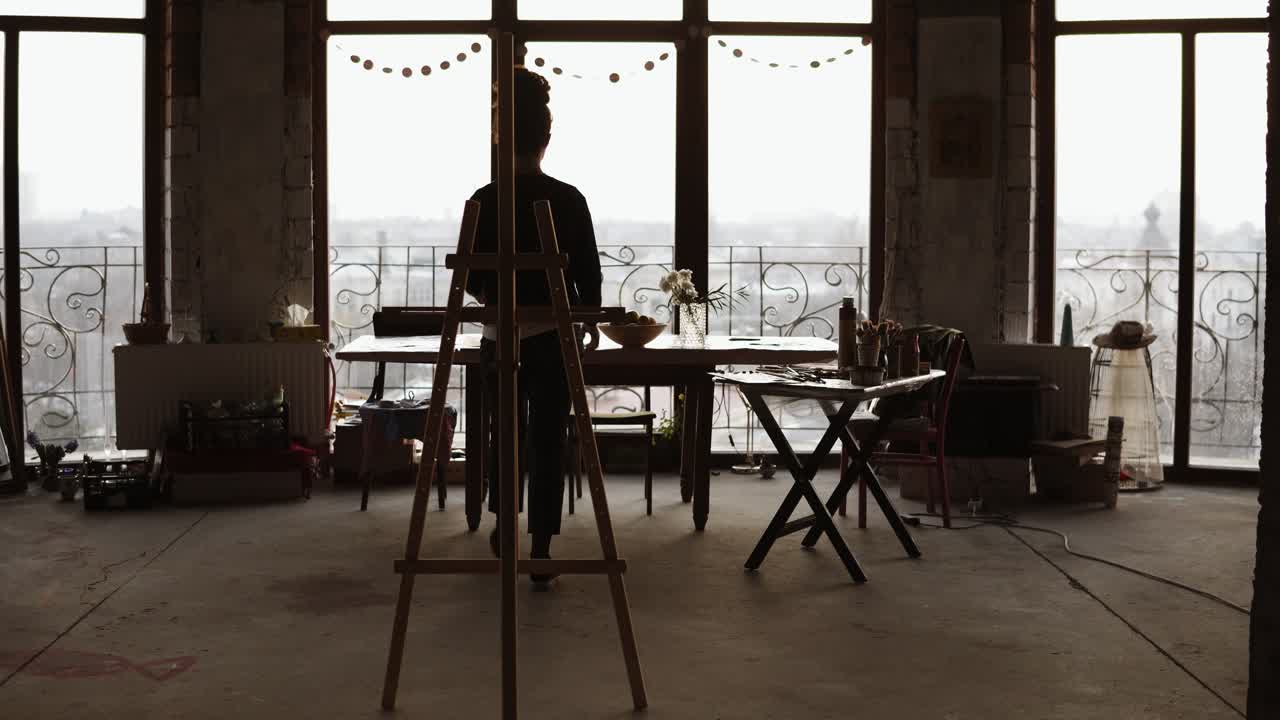 Artista donna, in controluce dalla luce del giorno proveniente dalle finestre panoramiche sullo sfondo, porta il cavalletto e vi posiziona la tela.
