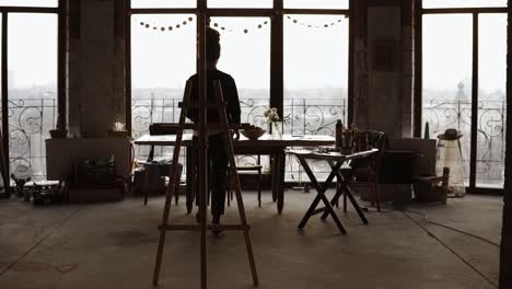 La-Artista-Femenina,-Sombreada-Por-La-Luz-Del-Día-En-El-Fondo-Que-Entra-Por-Las-Ventanas-Panorámicas,-Trae-El-Caballete-Y-Coloca-El-Lienzo-Sobre-él.