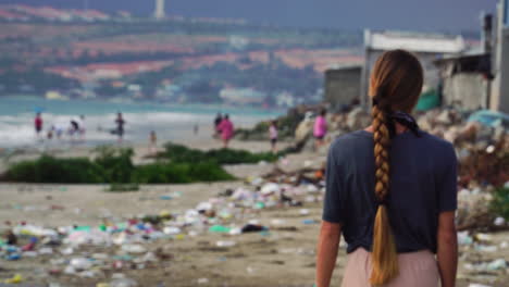 woman walking along sandy beach with empty plastic bottle and garbage spread across the beach