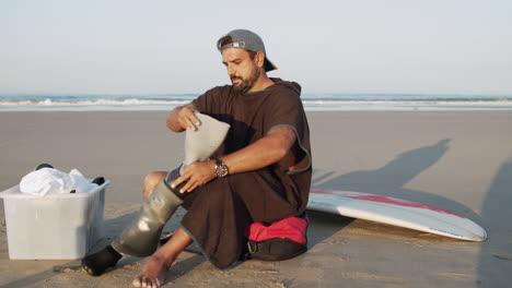 long shot of a male surfer sitting on ocean coast and putting prosthetic leg on