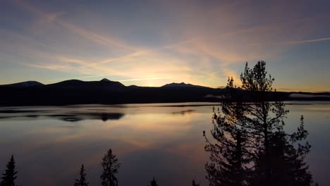 Reflective-lake-pull-back-through-silhouette-pine-trees-at-sunset,-60fps