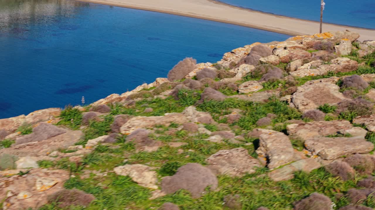 Angled aerial tracking along rocky coastline and blue water near Kythnos, dry vegetation and cliff detail visible to reveal Kolona Beach