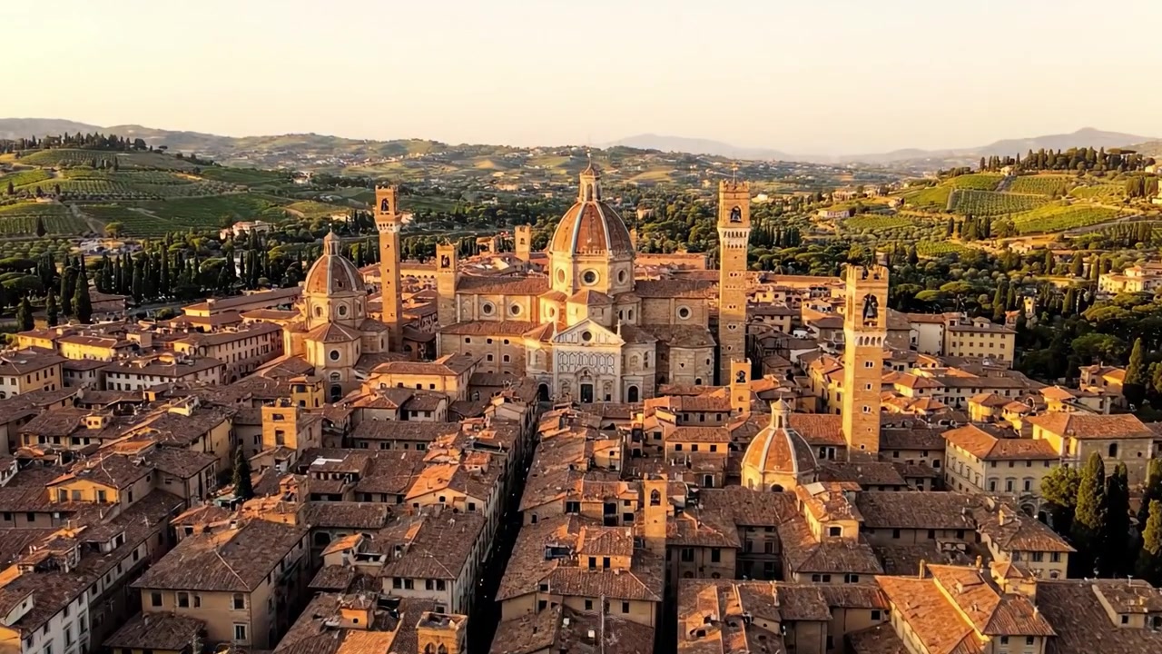 Vista aerea mozzafiato di una storica città italiana circondata da una vegetazione lussureggiante e dolci colline al tramonto, che ne evidenzia le meraviglie architettoniche.